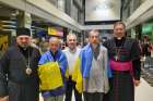 Draped in Ukrainian flags, Redemptorist Fathers Bohdan Geleta, left, and Ivan Levytsky, right, pose for a photo at the Kyiv airport in Ukraine June 29, 2024. To the far right is Archbishop Visvaldas Kulbokas, apostolic nuncio to Ukraine.